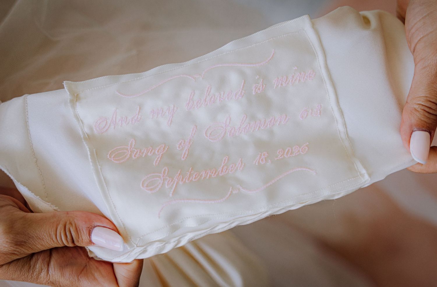 Close-up of a wedding dress with embroidered pink cursive text on white satin fabric held by a bride.