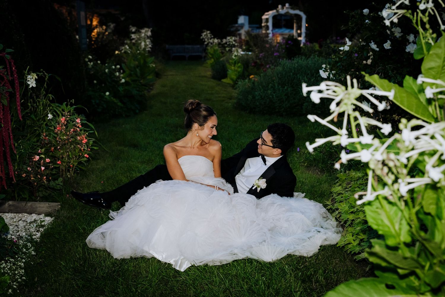 Bride and groom sitting together in a lush garden at night, sharing a romantic moment in wedding attire.