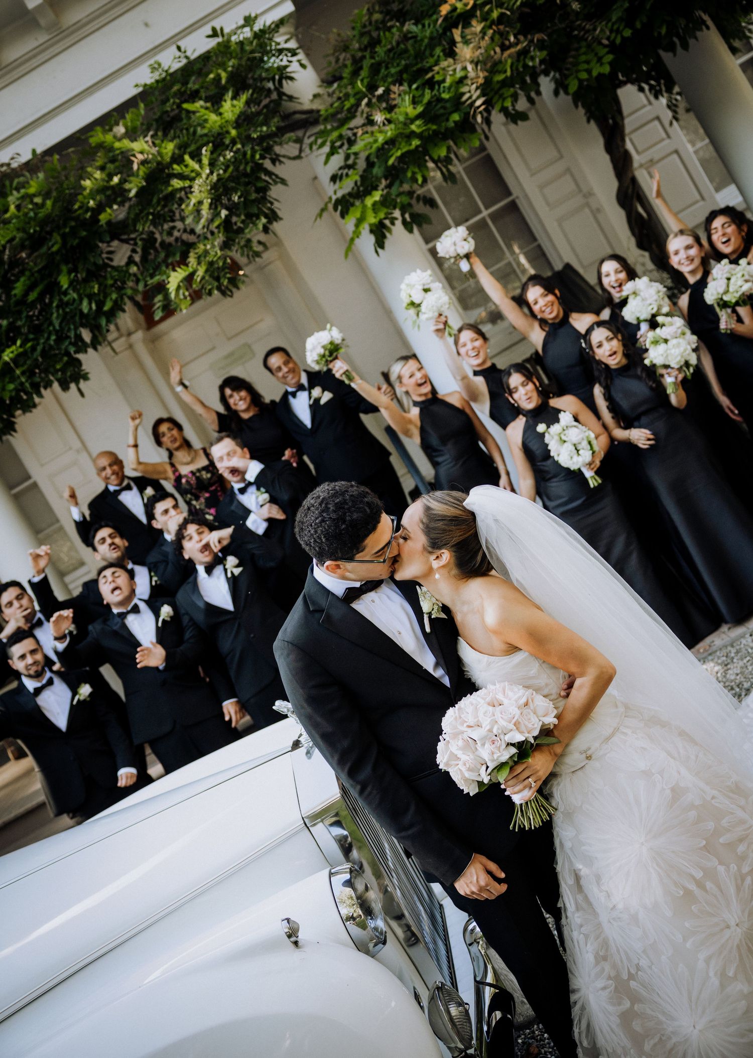 Bride and groom share a kiss in front of wedding party dressed in black tuxedos and gowns holding white bouquets.