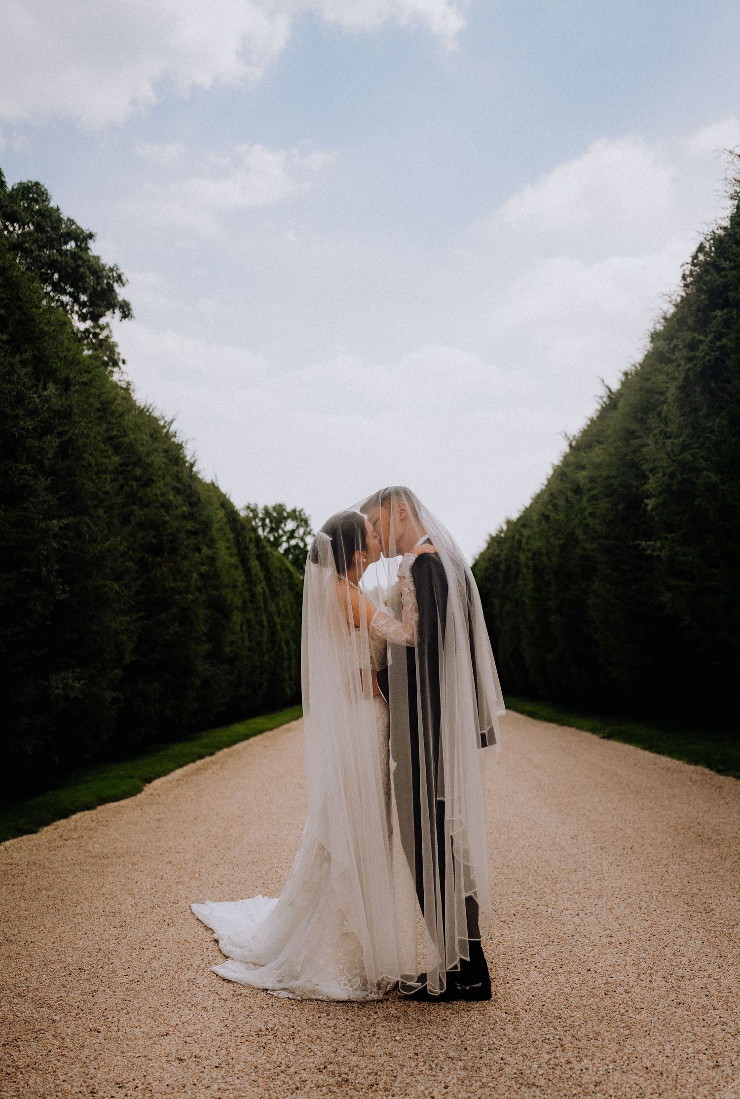 Bride and groom share a kiss under a veil on a gravel path lined with tall hedges at an elegant outdoor wedding.