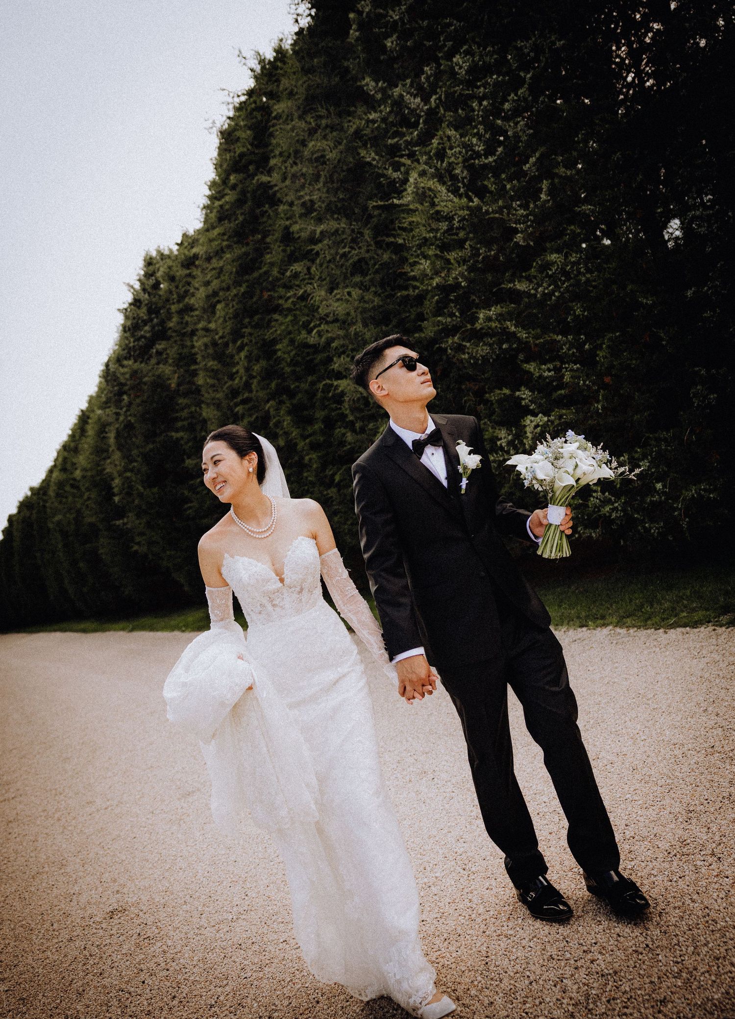 Bride in white lace gown and groom in black tuxedo hold hands on gravel path with tall hedges backdrop.