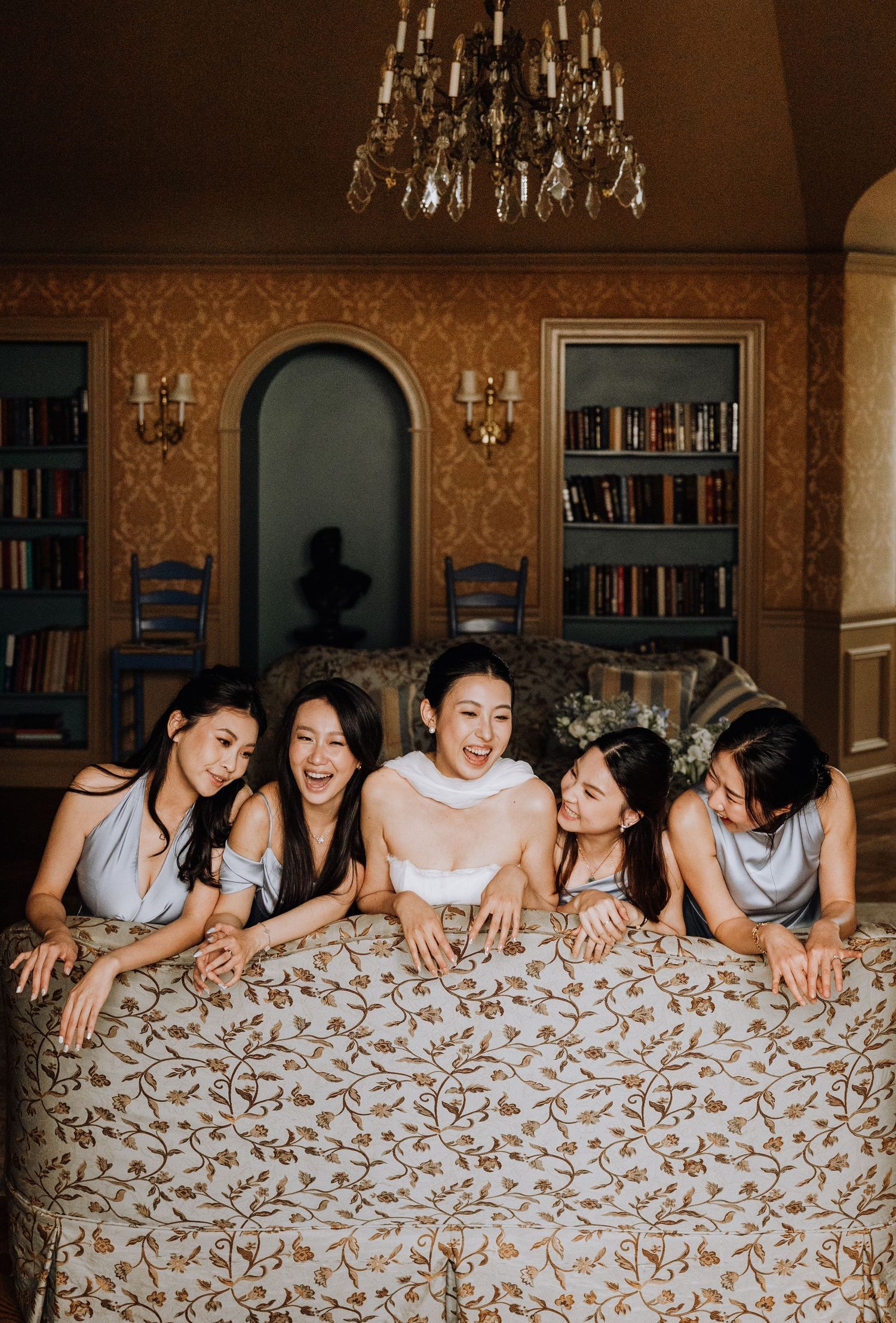 Five bridesmaids in silver dresses lean over an ornate bed laughing with bride in elegant chandelier-lit room.