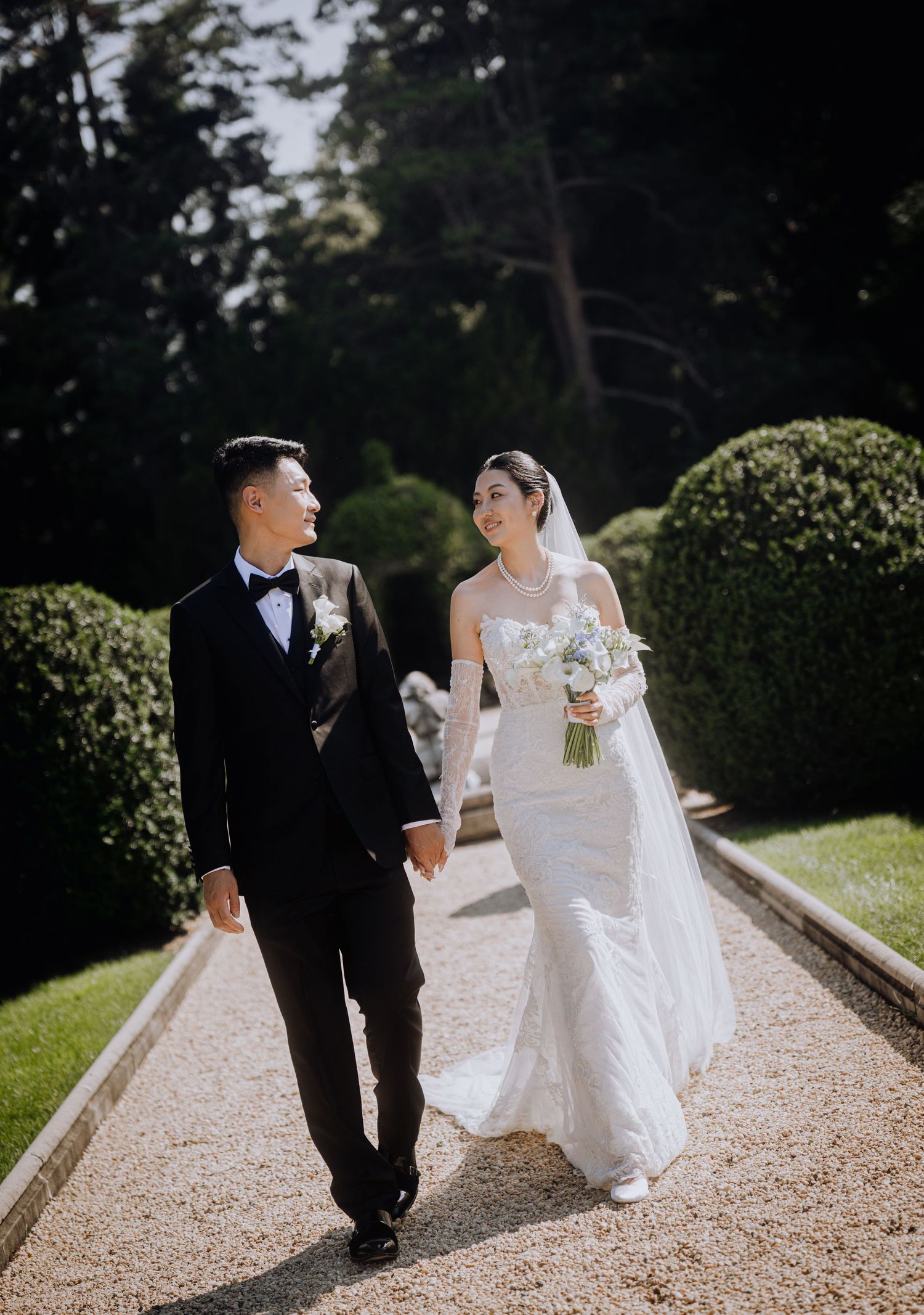Bride and groom walk hand in hand through elegant garden path, bride in white off-shoulder gown holding bouquet.