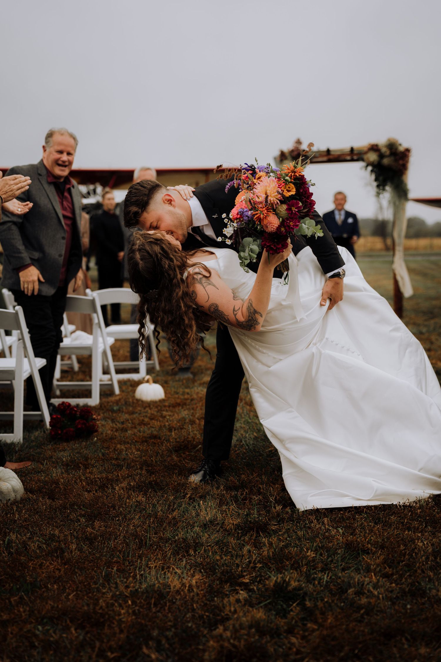 Joyful wedding dip pose with colorful bouquet and guests celebrating in background.