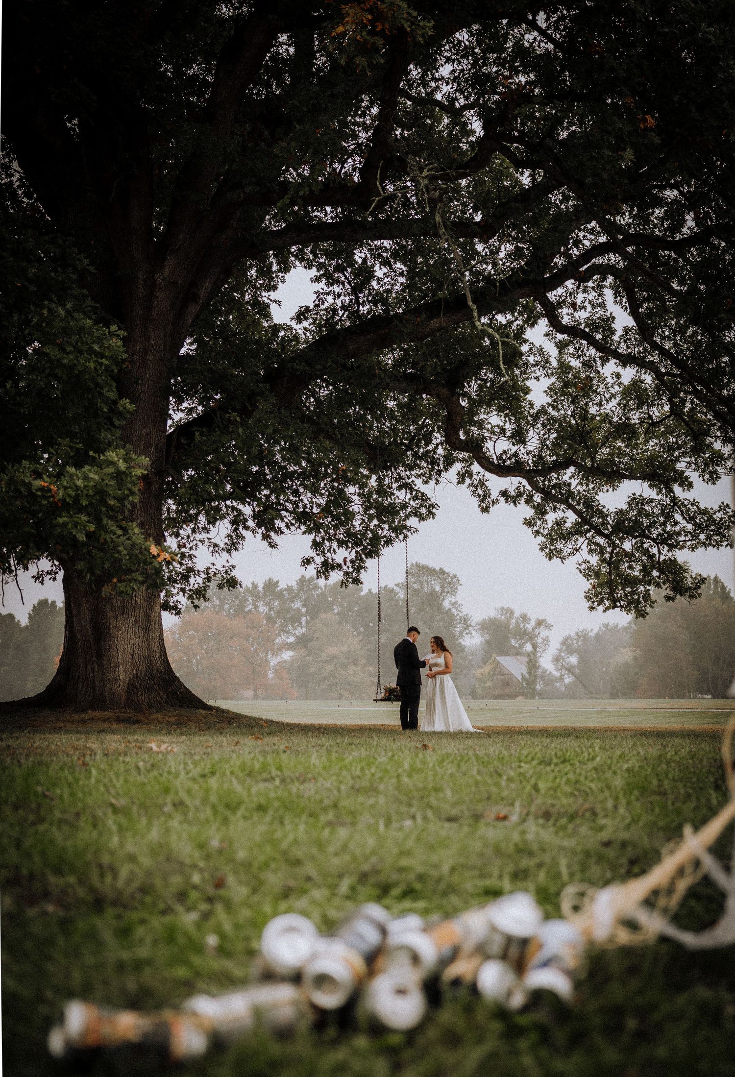 Wedding string lights frame a distant couple under large oak trees in a misty field.