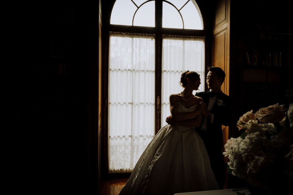 Romantic silhouette of bride and groom embracing by an elegant arched window in a dimly lit room.