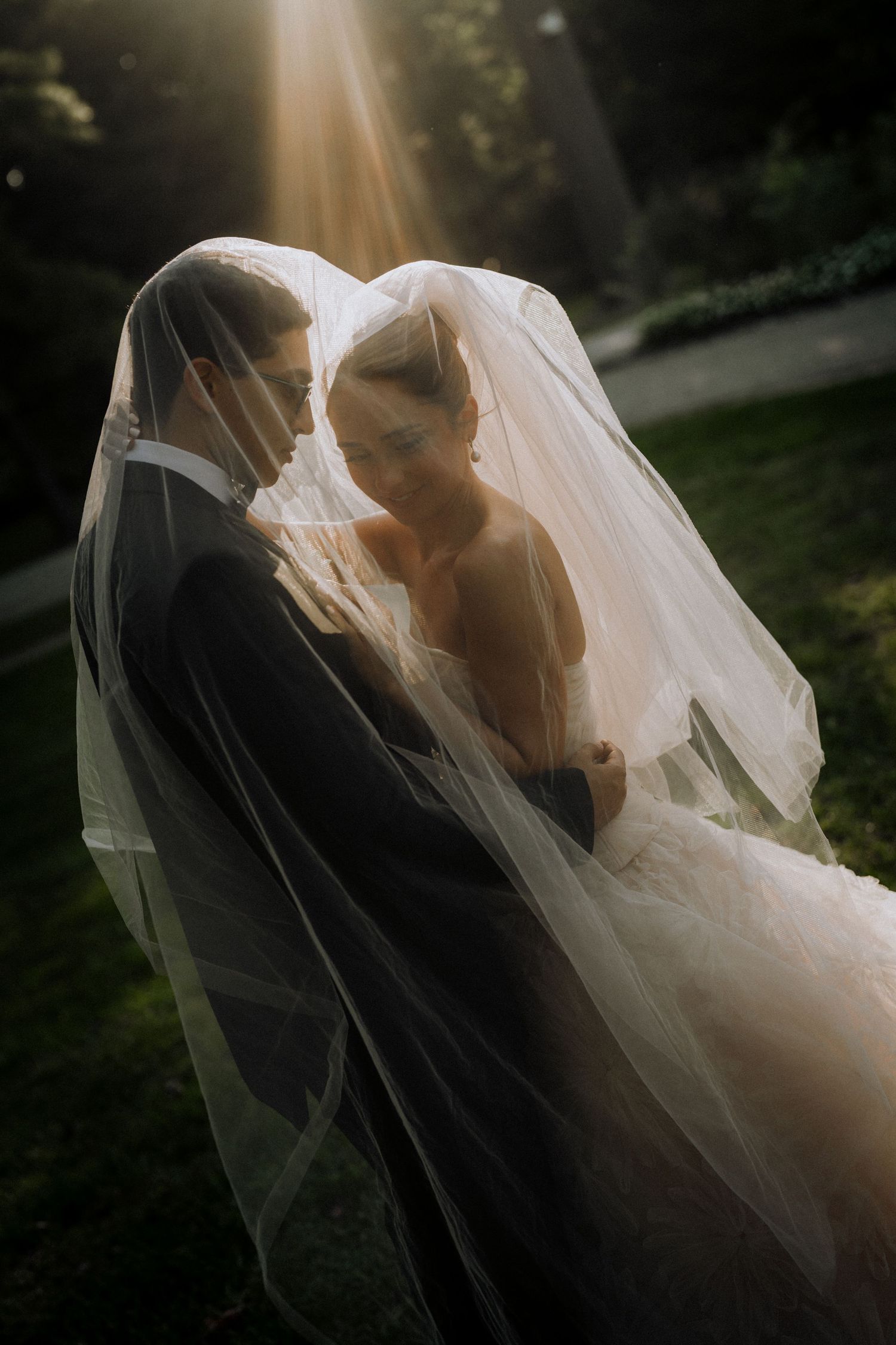 Bride and groom share an intimate moment under a flowing veil, bathed in golden sunlight in a lush garden.