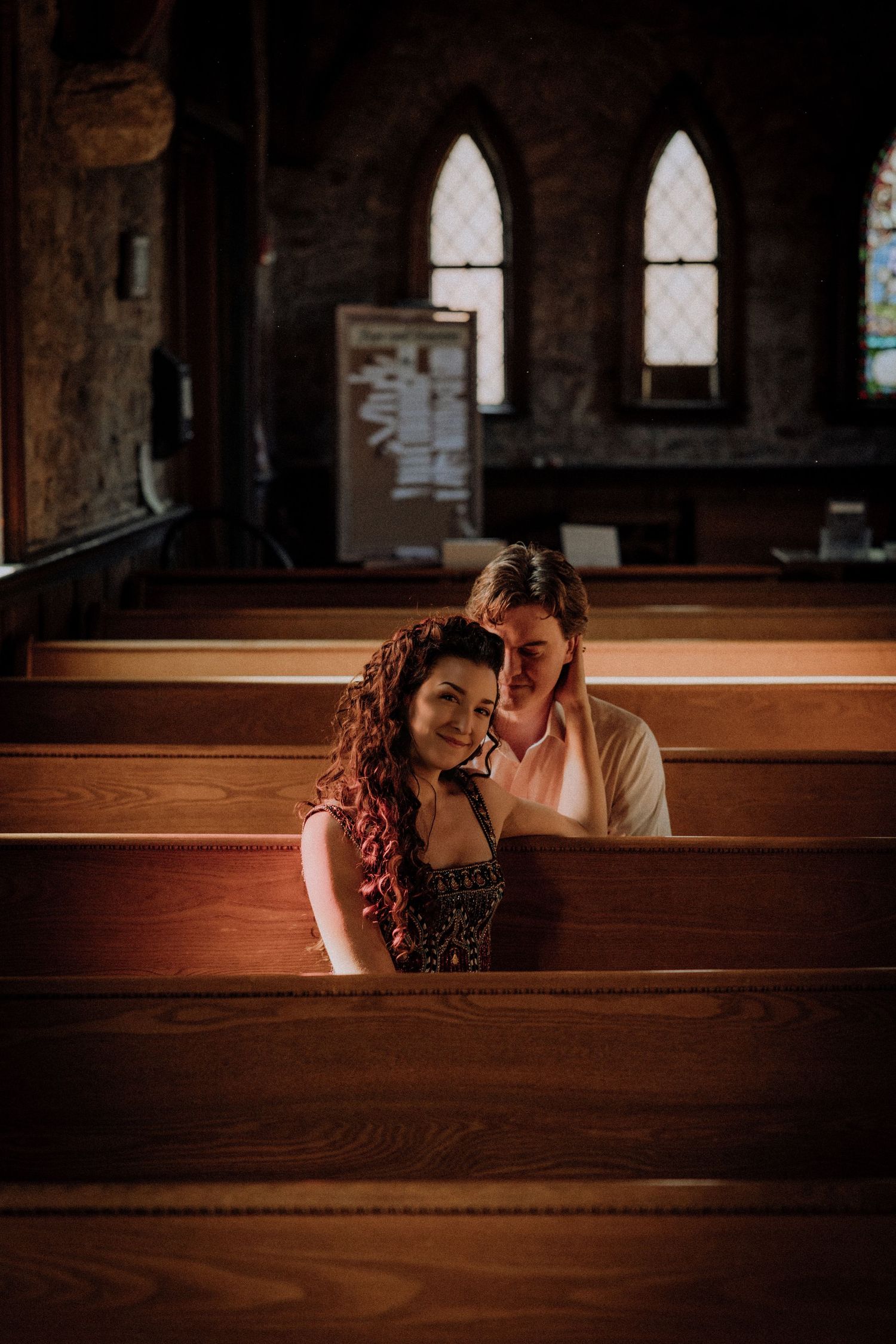 Couple sharing tender moment while sitting in wooden church pew with arched windows in background.