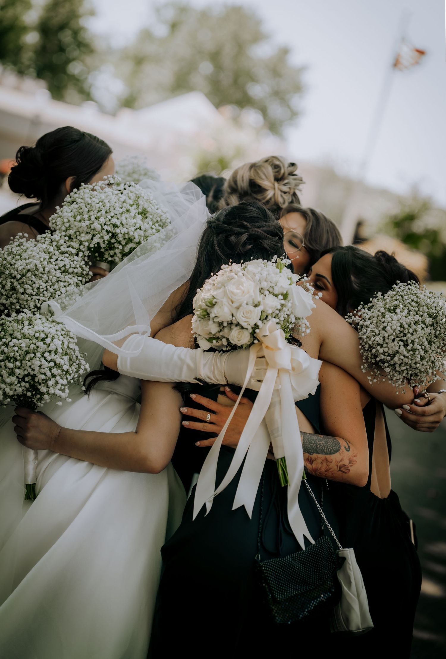 Bridesmaids in black dresses embrace while holding white baby's breath bouquets.