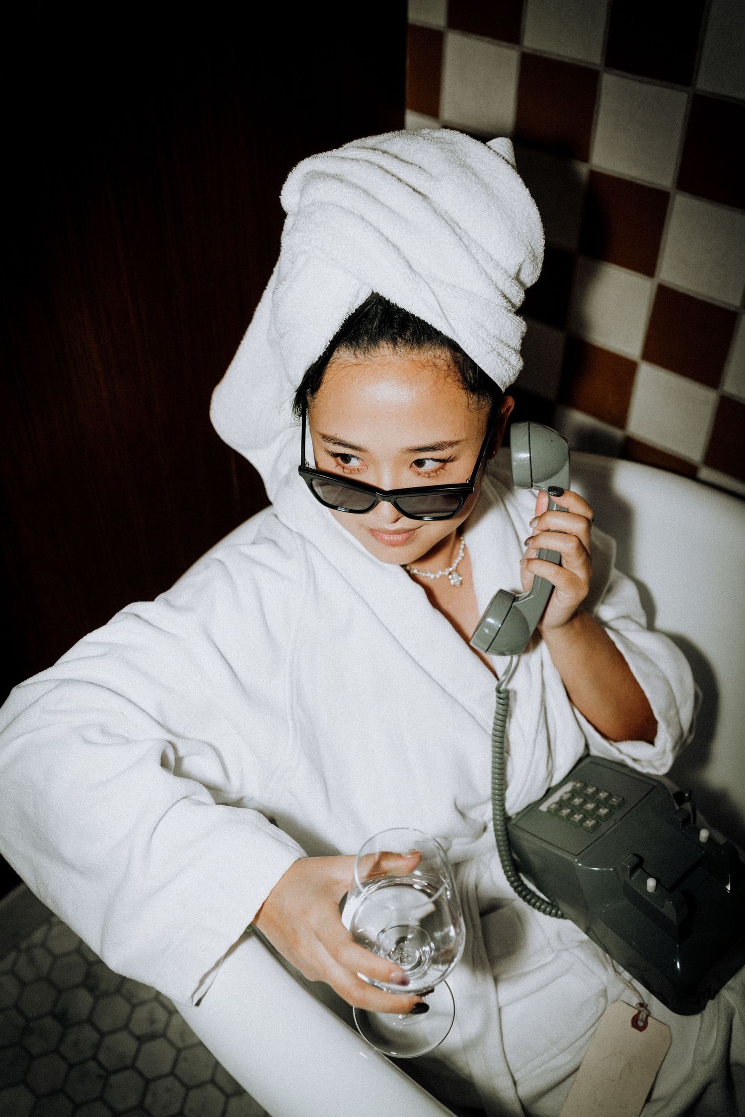 Woman in white bathrobe and towel turban, wearing sunglasses, holding retro phone and glass of water in bathroom.