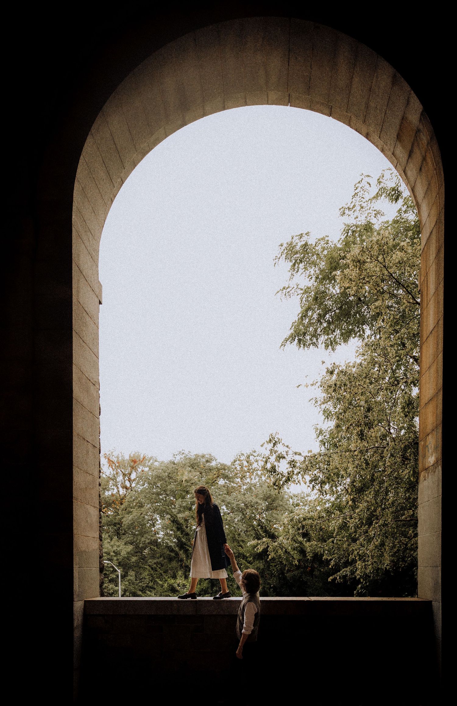A silhouetted figure sits in an arched stone window frame overlooking trees and greenery.