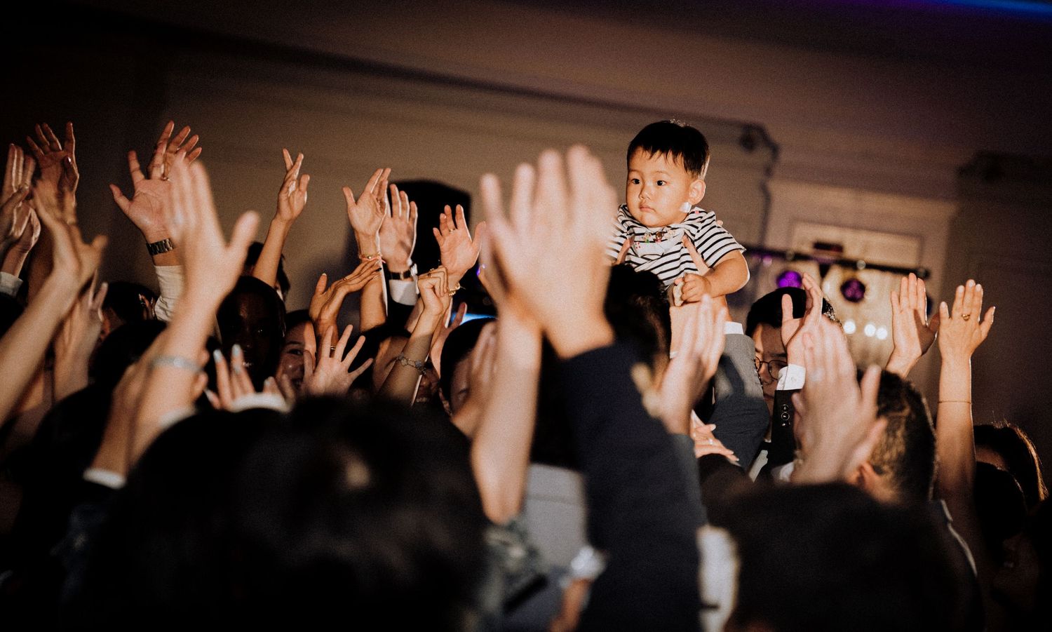 Young child raised above crowd with hands in air at lively Asian event or concert celebration.