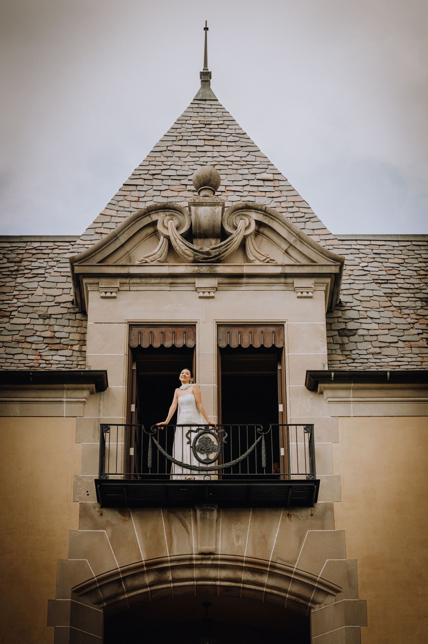 Bride in white gown standing on ornate stone balcony of elegant historic mansion with slate roof.
