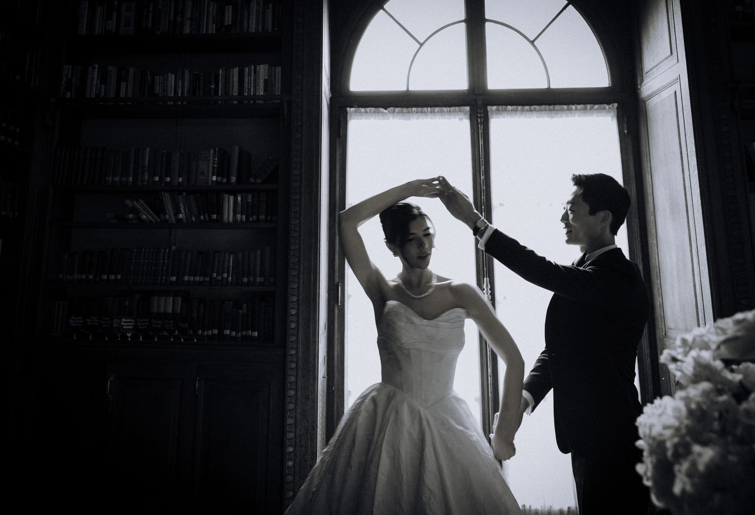 Bride and groom dancing elegantly in front of a grand window in a dimly lit library, black and white wedding photo.