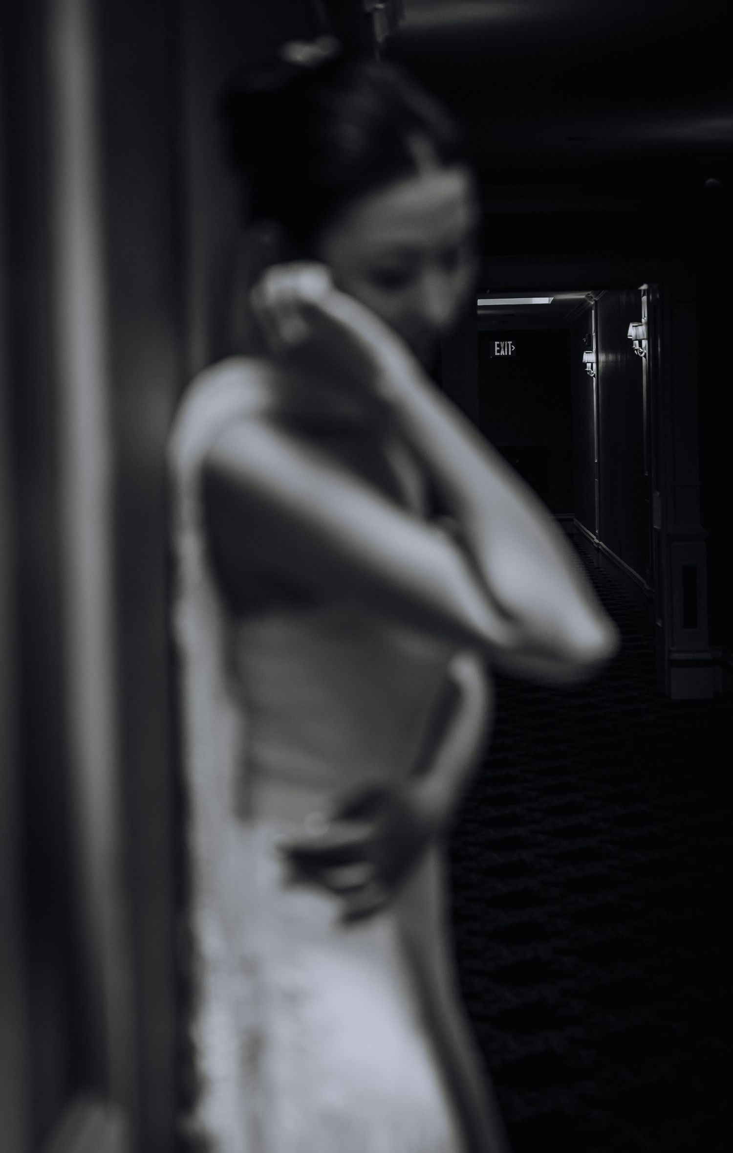 Black and white photo of a woman in a white dress standing in a dimly lit hallway, looking down pensively.