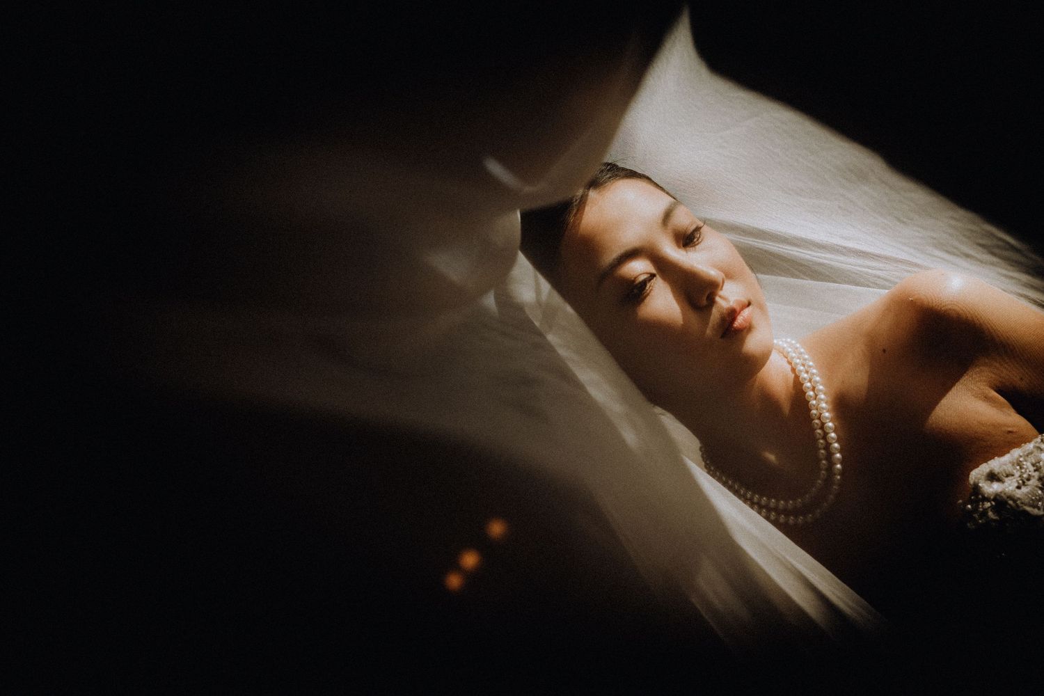 A bride in pearl necklace and lace gown gazes upward under a flowing white veil in a dramatic dark setting.