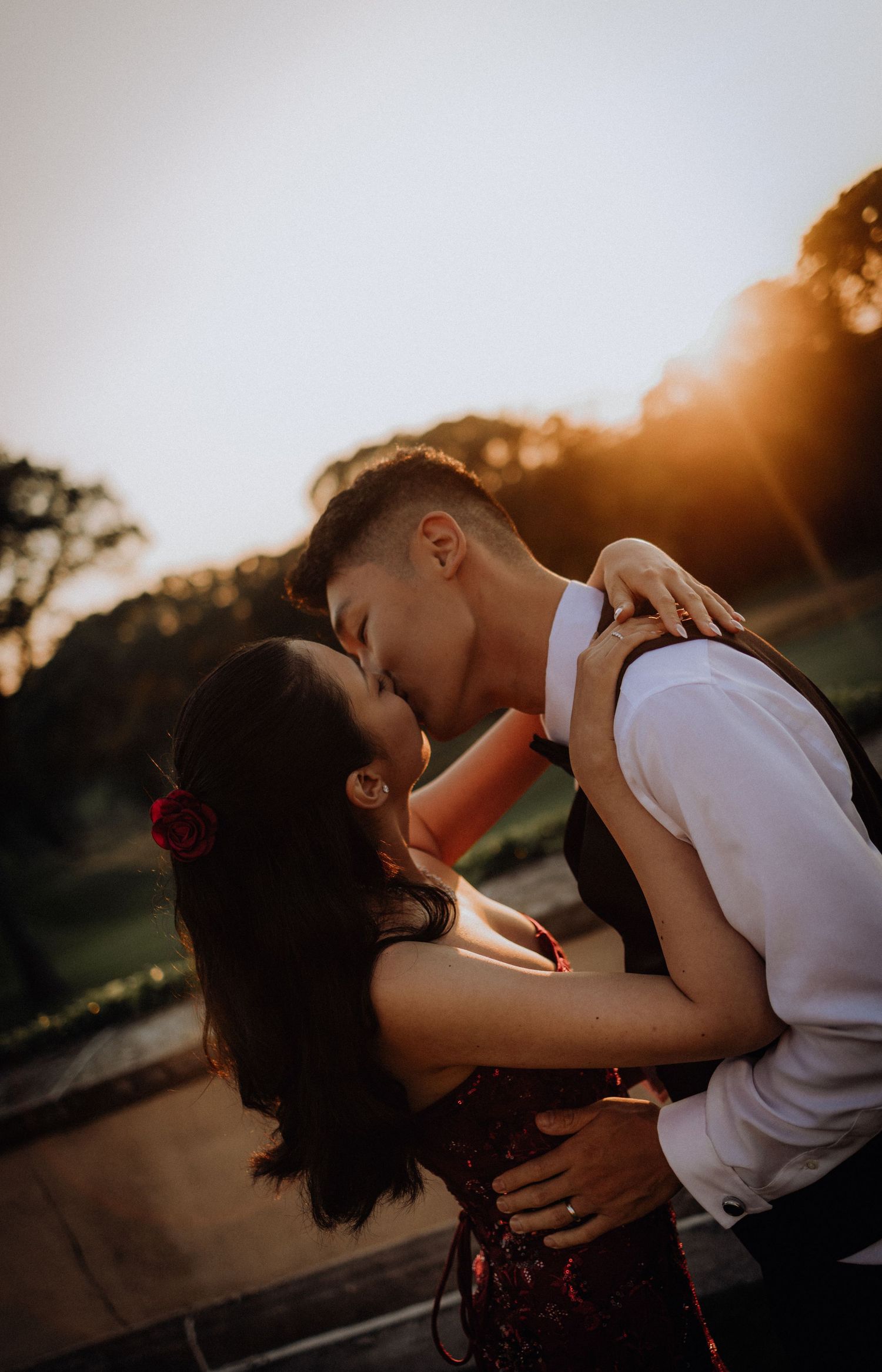 Couple sharing a romantic kiss at golden hour sunset, woman wearing red floral dress with rose hair accessory outdoors.