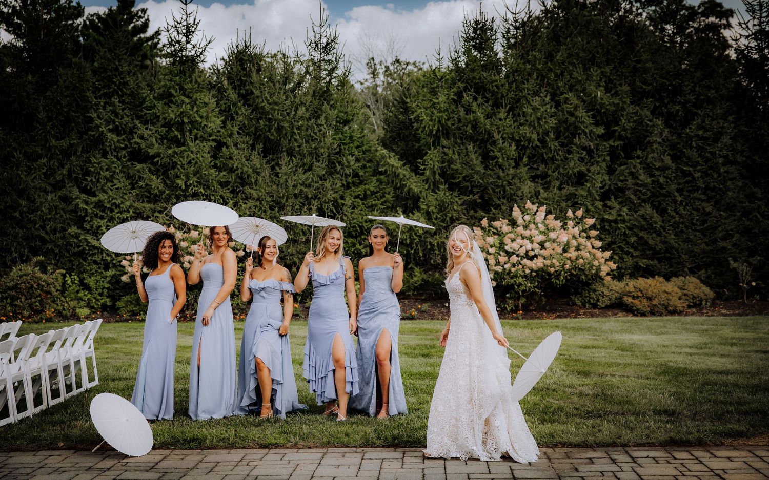 Bridal party in pale blue dresses poses with parasols on a garden pathway.