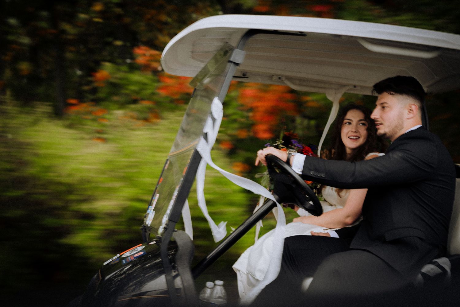 Newlyweds enjoying a ride in a golf cart through autumn scenery.