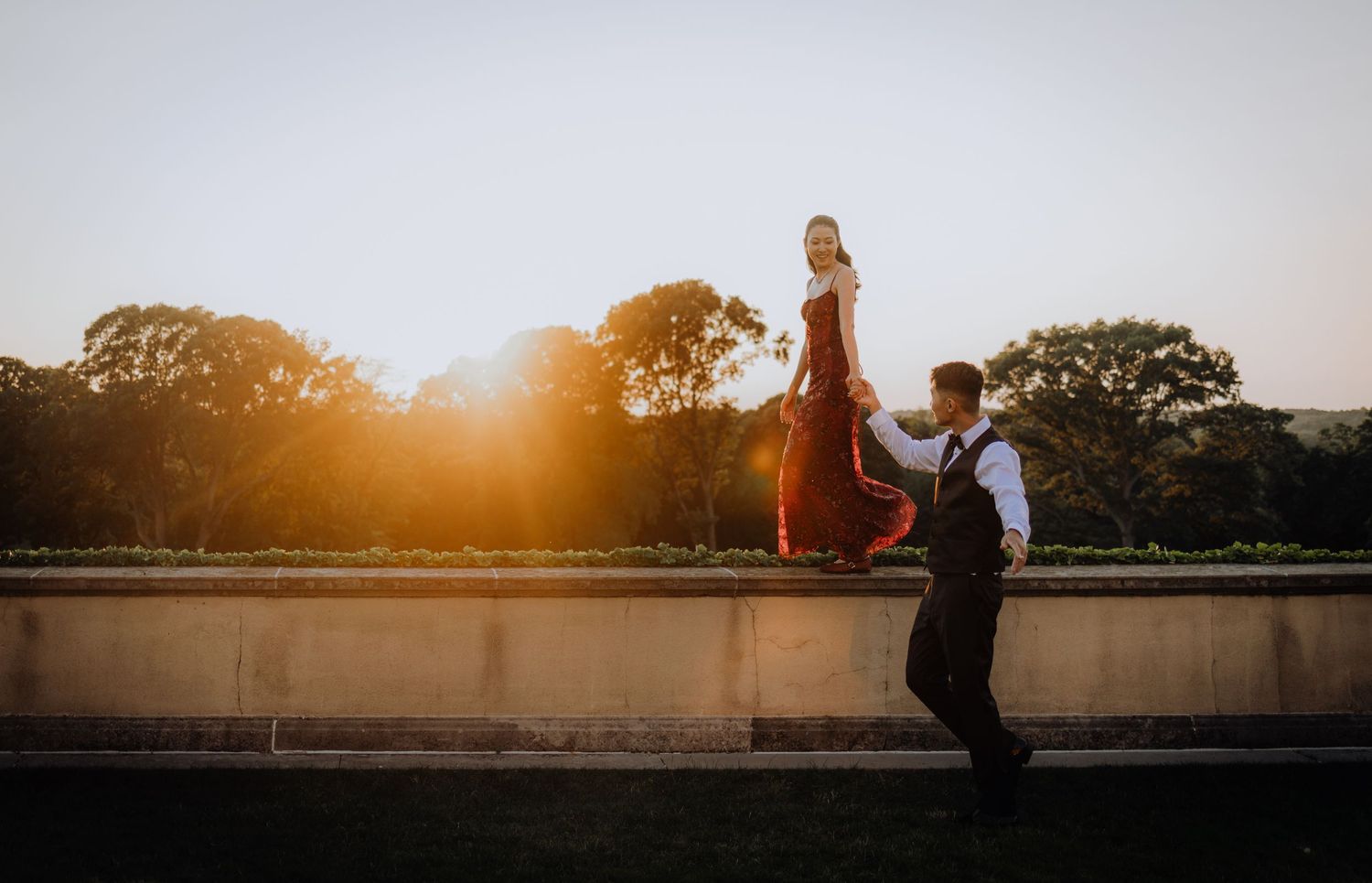 Couple in golden sunset light, woman in red gown walks wall as man follows below, romantic outdoor engagement photo.