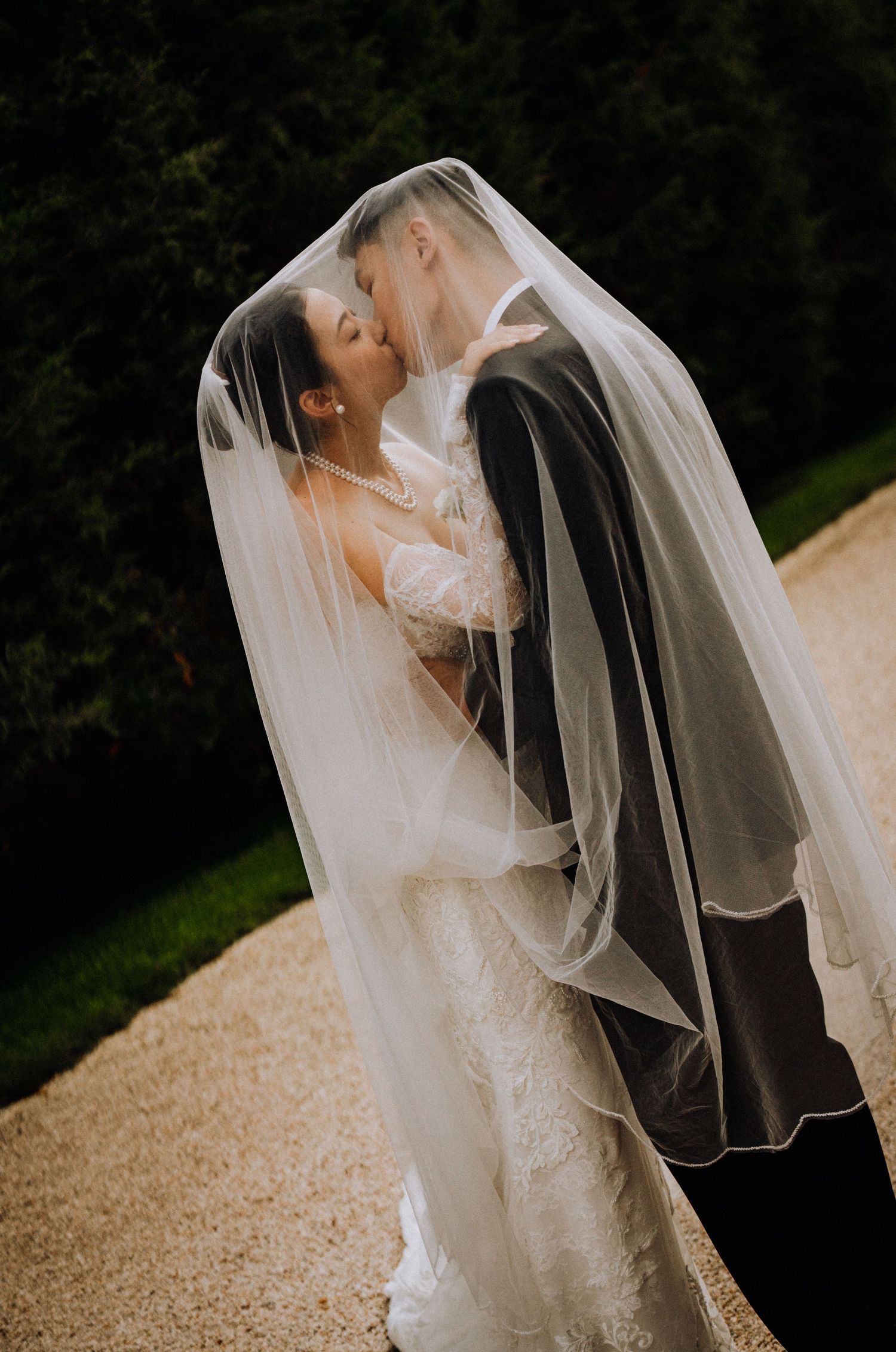Bride and groom sharing a romantic kiss under a flowing white veil outdoors on their wedding day.