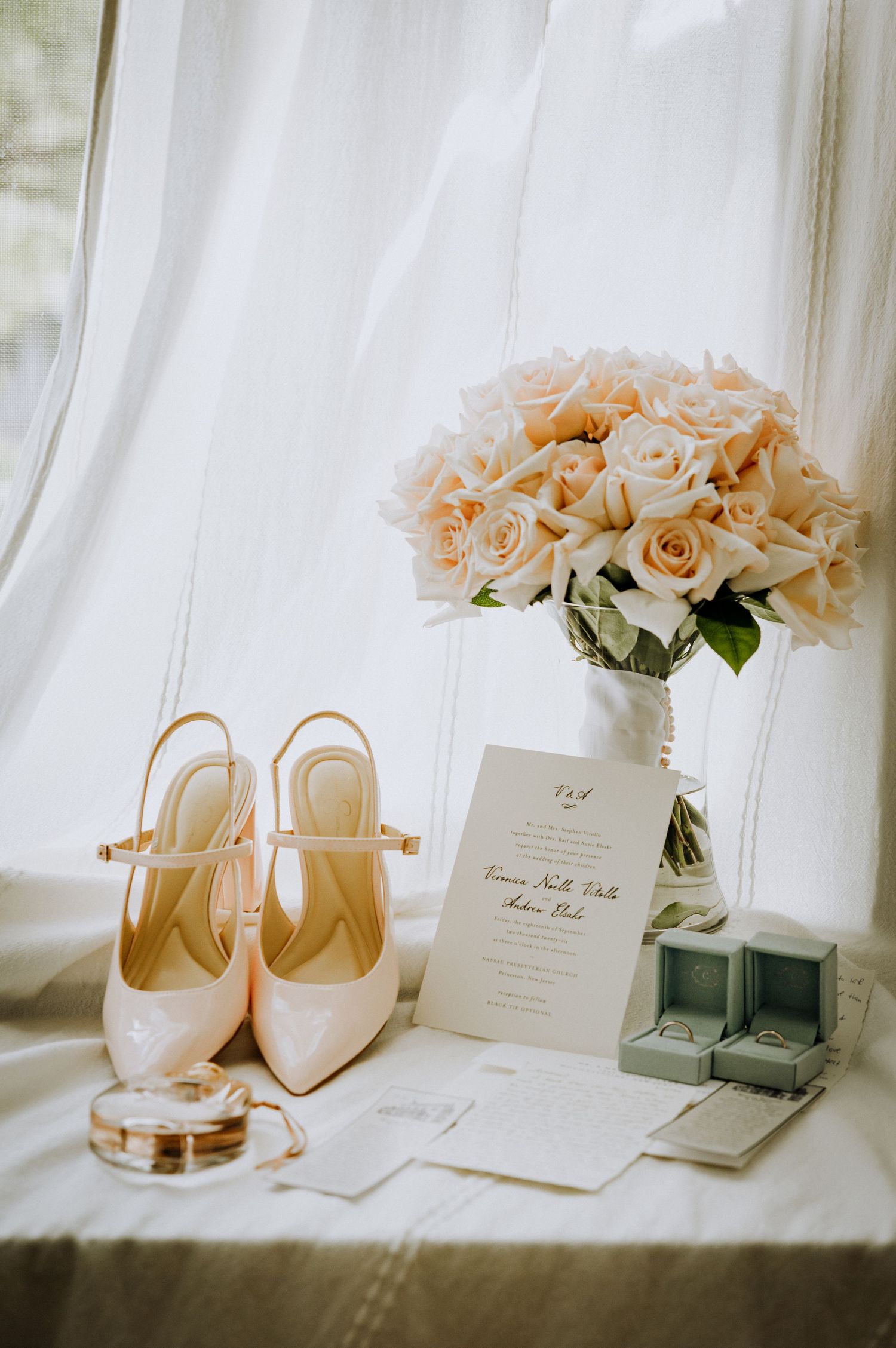 Elegant wedding flat lay featuring blush heels, pink roses bouquet, invitation suite, and ring boxes on a soft white surface.