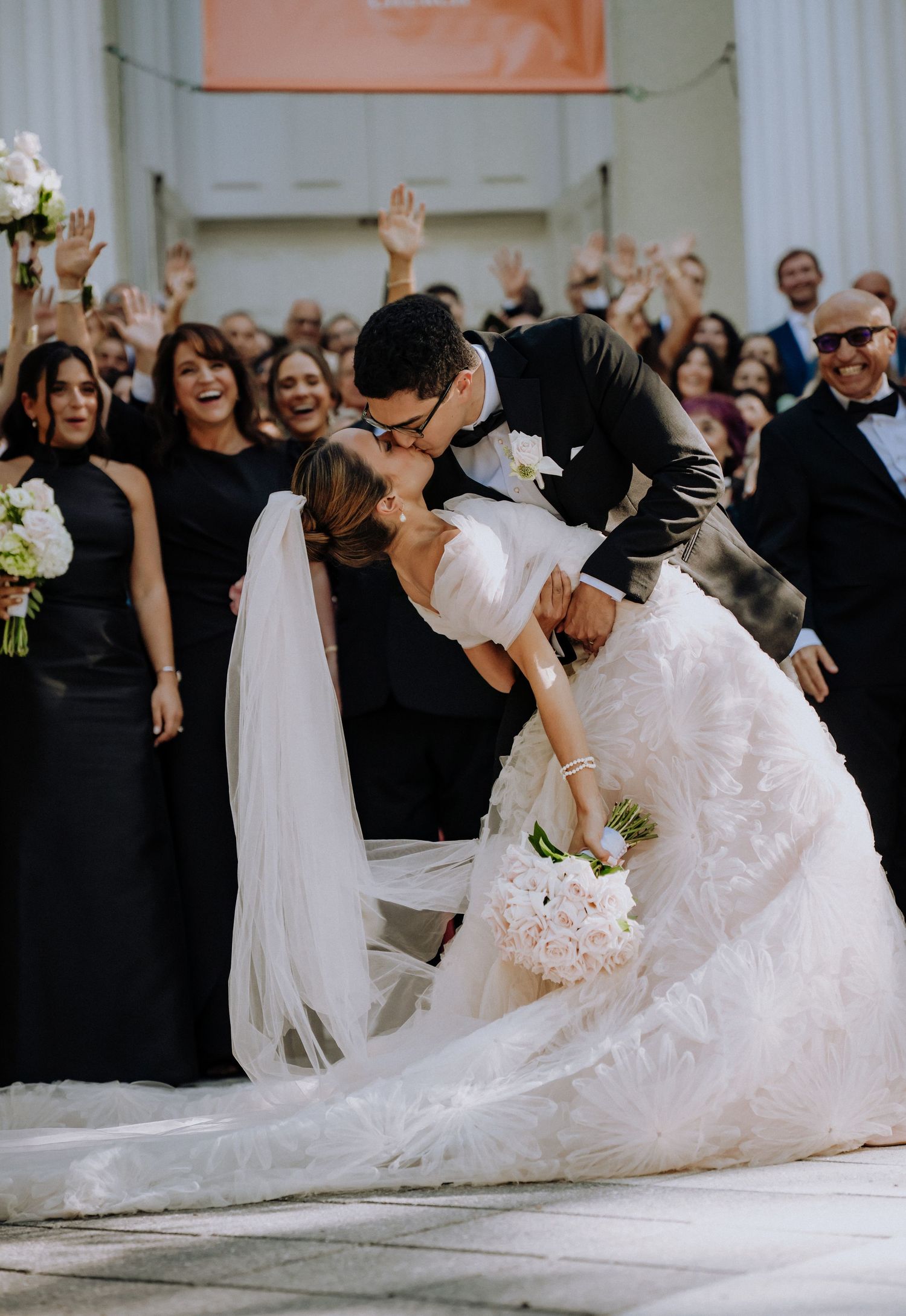 Groom dips bride in stunning white gown for a romantic kiss, surrounded by cheering guests in black attire.