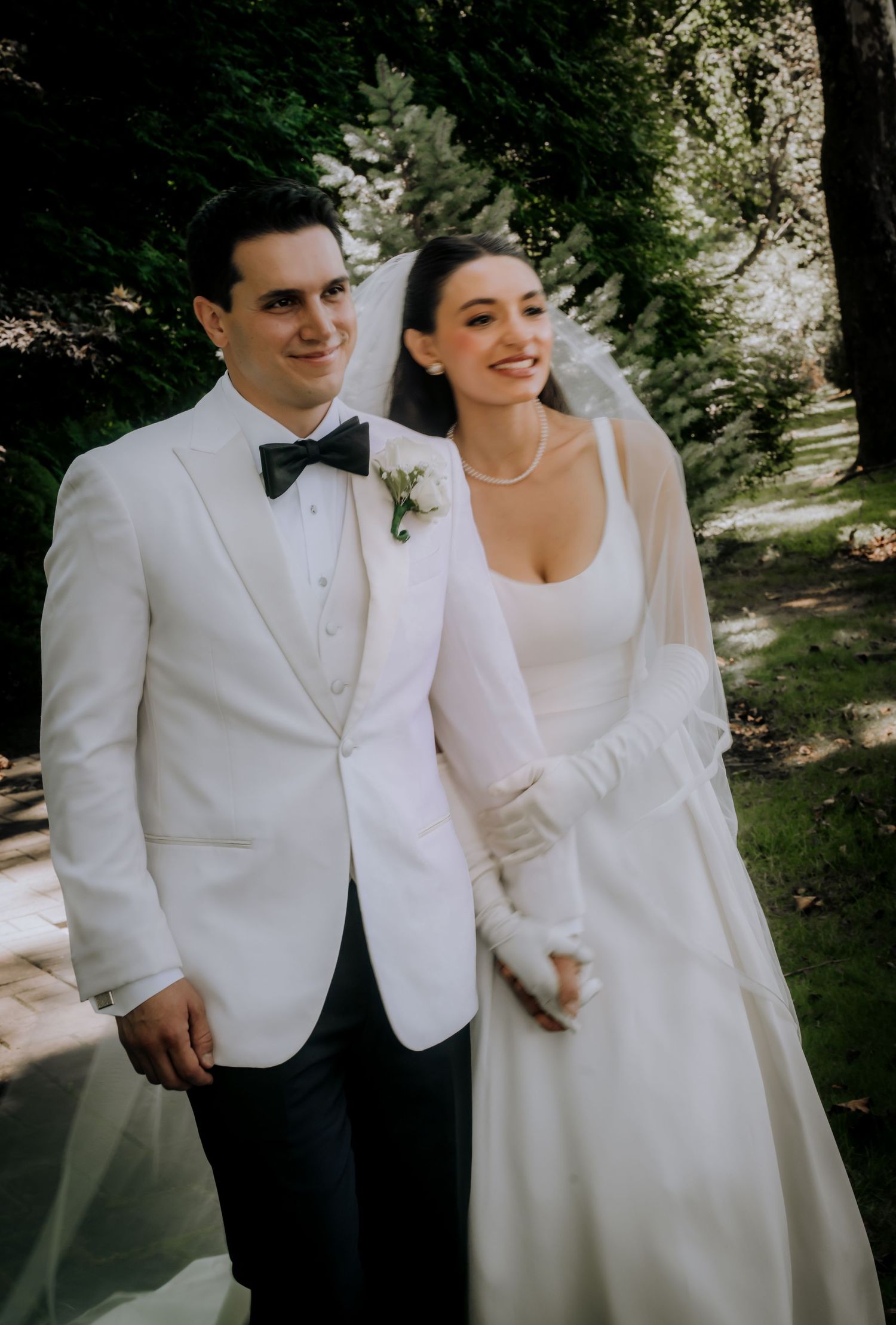 Elegant bride and groom in white tuxedo and wedding gown with veil smiling outdoors among lush green trees.Editorial and Cinematic Wedding p