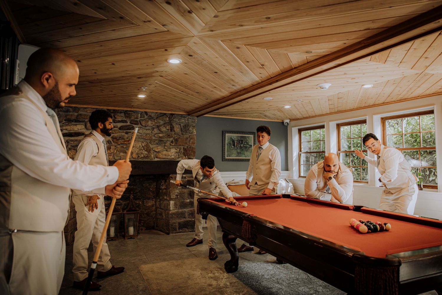 Groomsmen in light colored suits playing pool in a rustic room with wooden ceiling.Editorial and Cinematic Wedding photos
