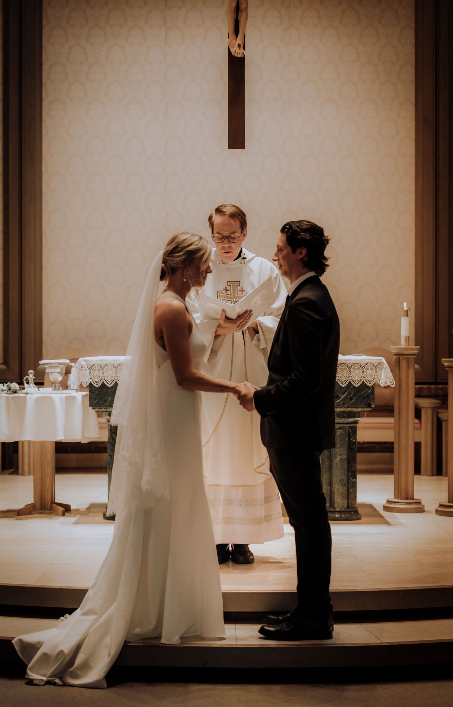 A wedding ceremony takes place at the altar with a priest officiating under a cross.