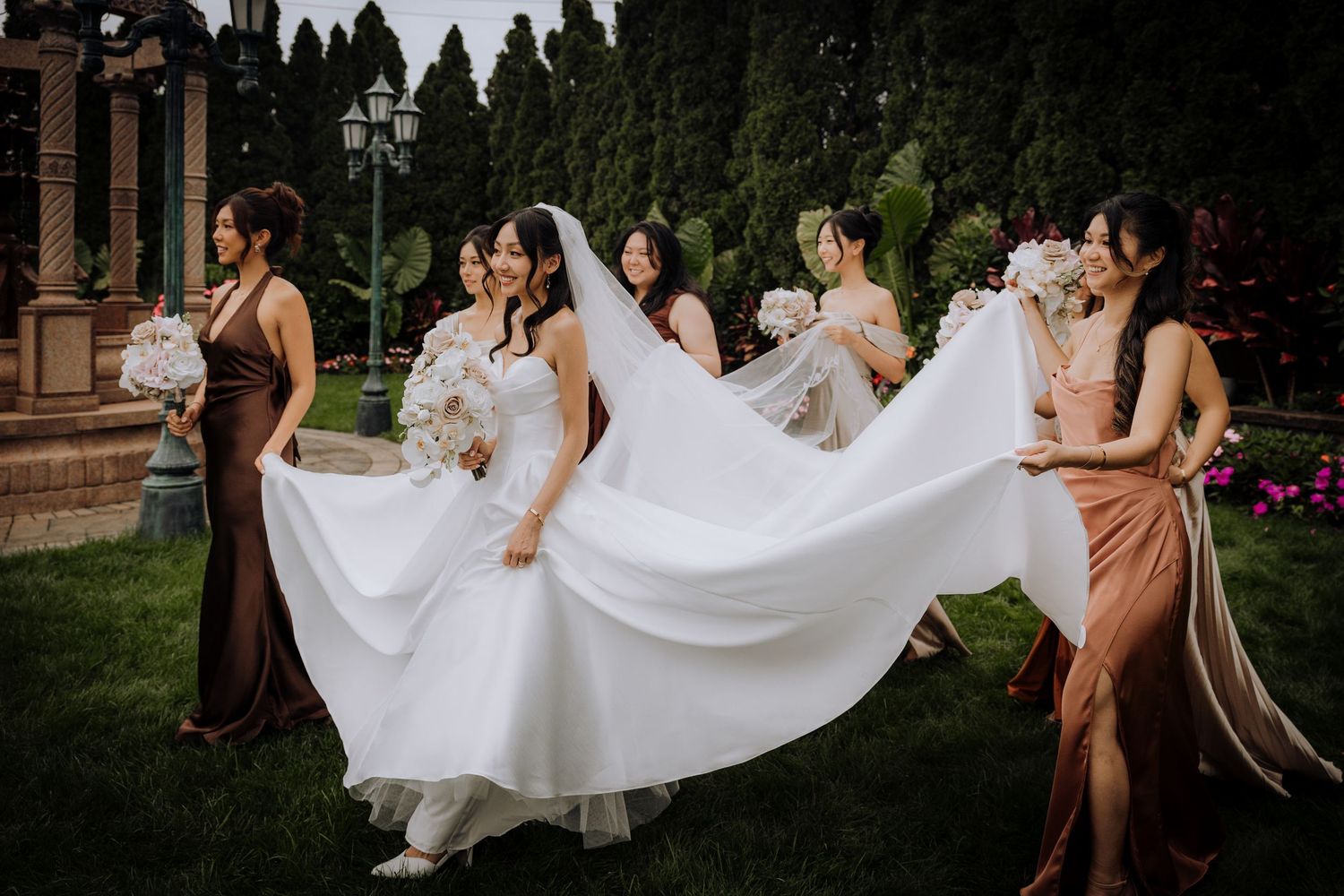 Wedding party arranges bride's flowing dress on garden lawn.
