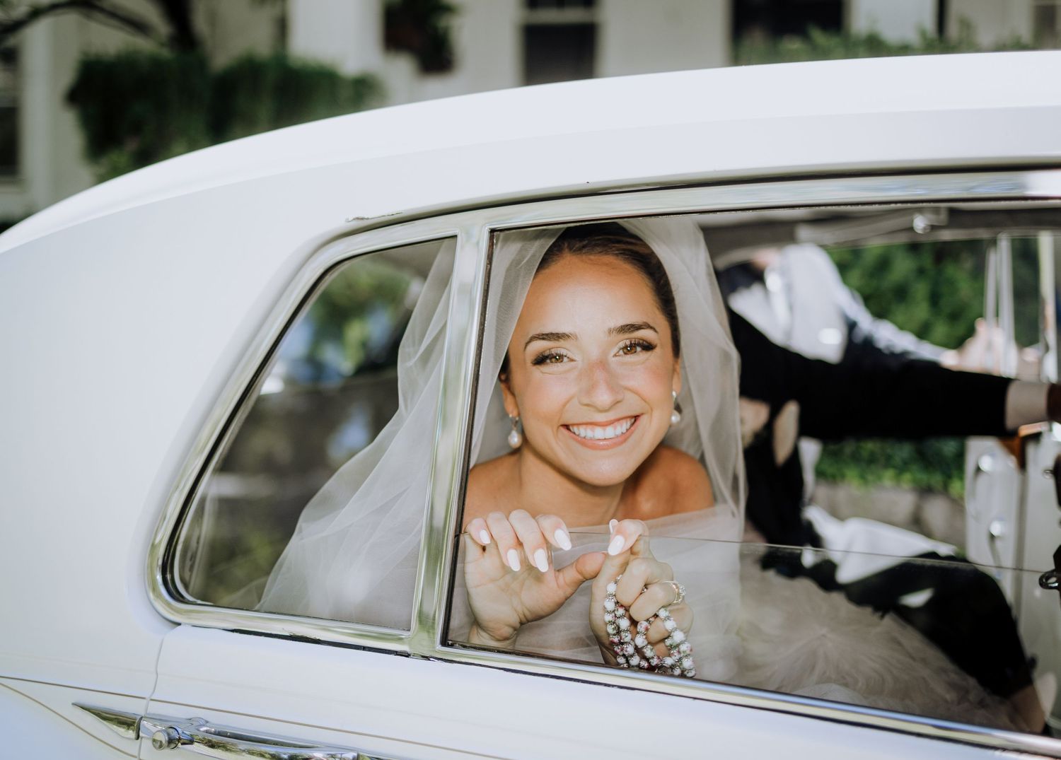 Smiling bride in a white veil sitting in a classic white car on her wedding day.