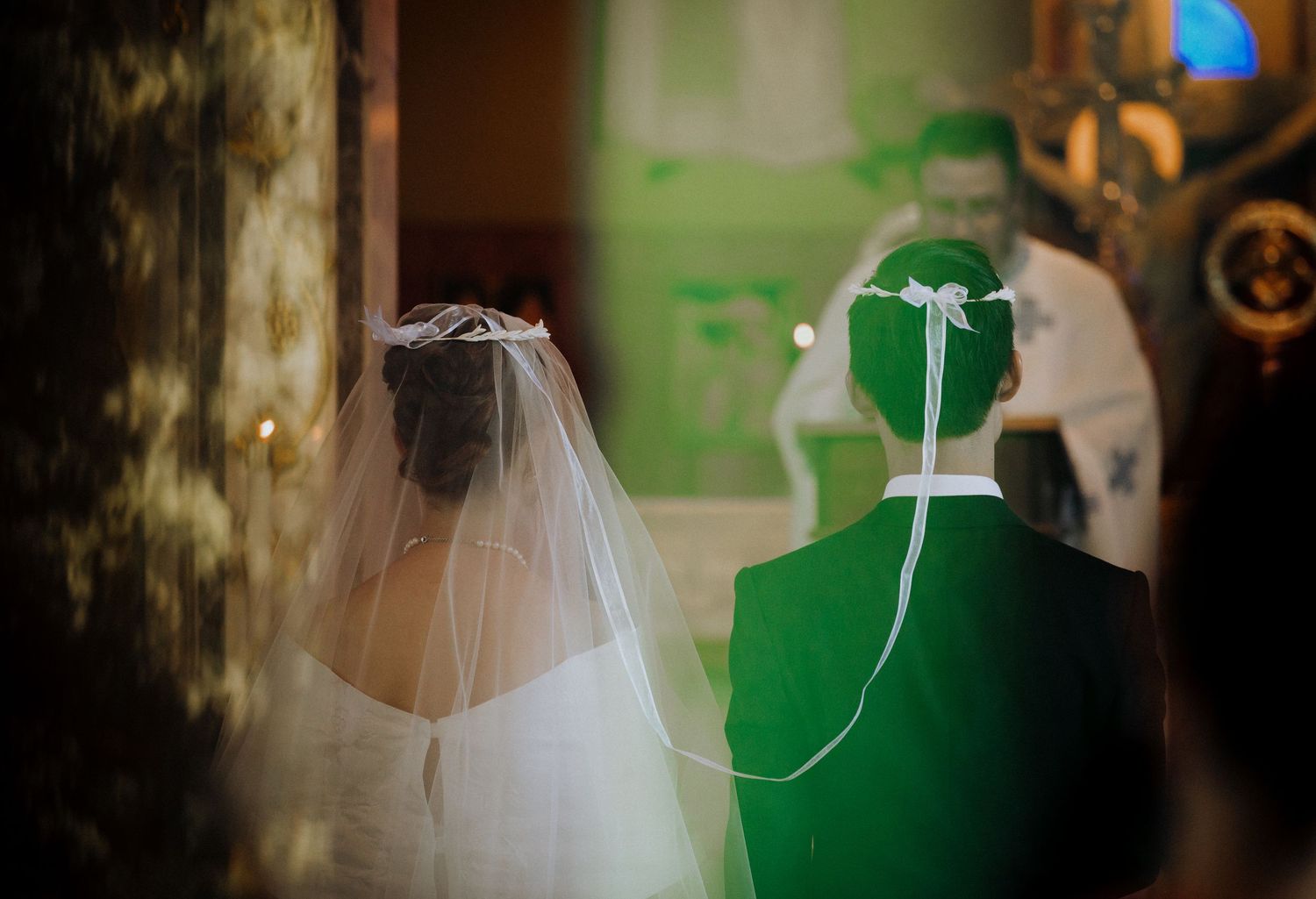Bride in white veil and groom in green suit stand before officiant during wedding ceremony.