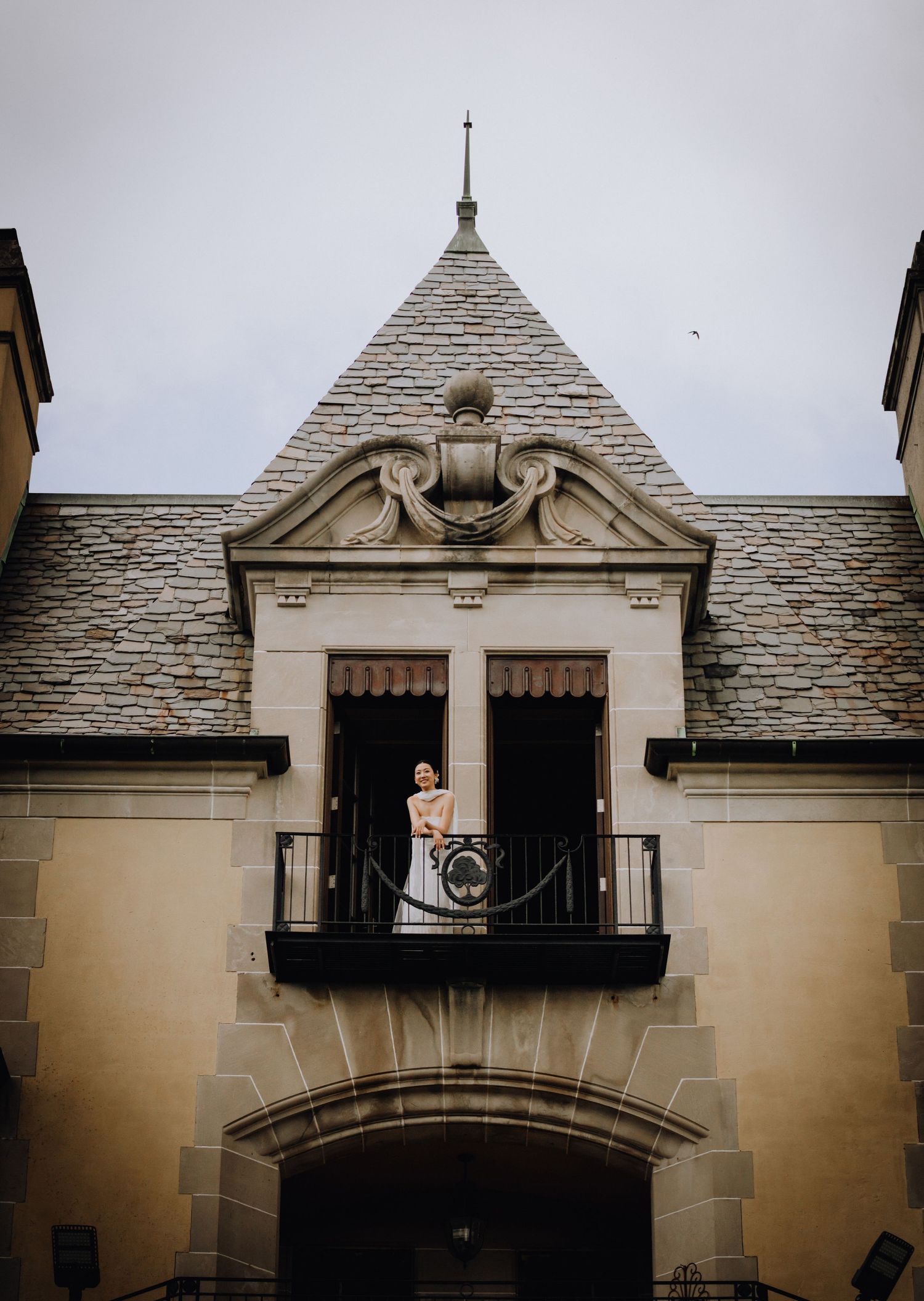 Bride in wedding dress standing on ornate castle balcony with slate roof and decorative stonework under overcast sky.