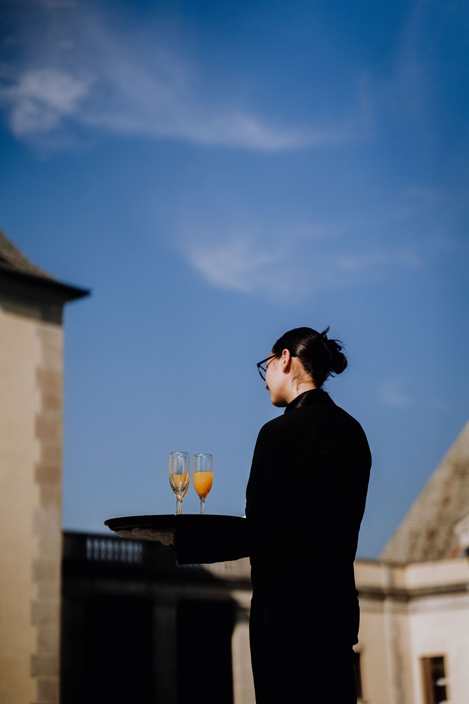 A waiter in black uniform holds a tray with two champagne flutes of orange juice against a blue sky.