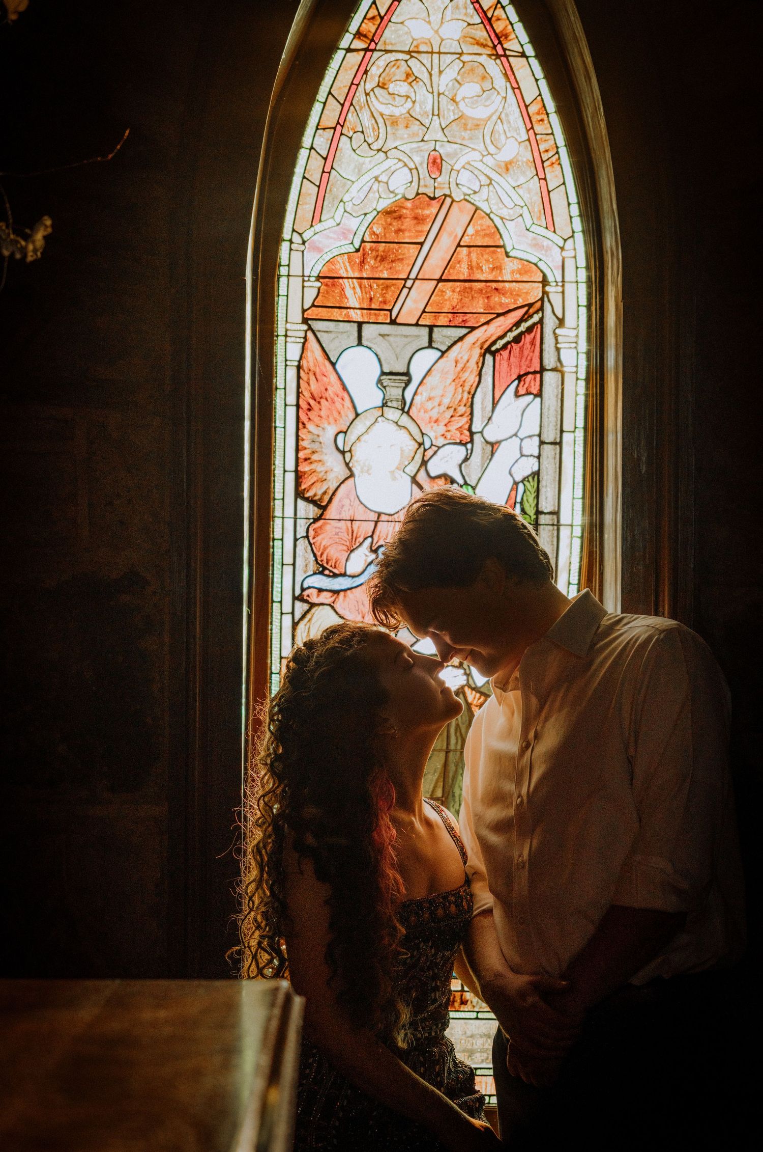 Couple embracing in front of ornate stained glass window with warm sunlight streaming through.
