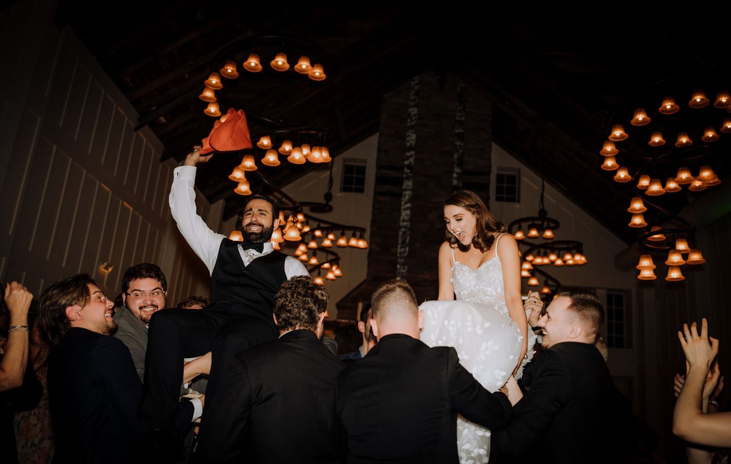 Wedding guests lift the newlyweds during a lively reception celebration under string lights.