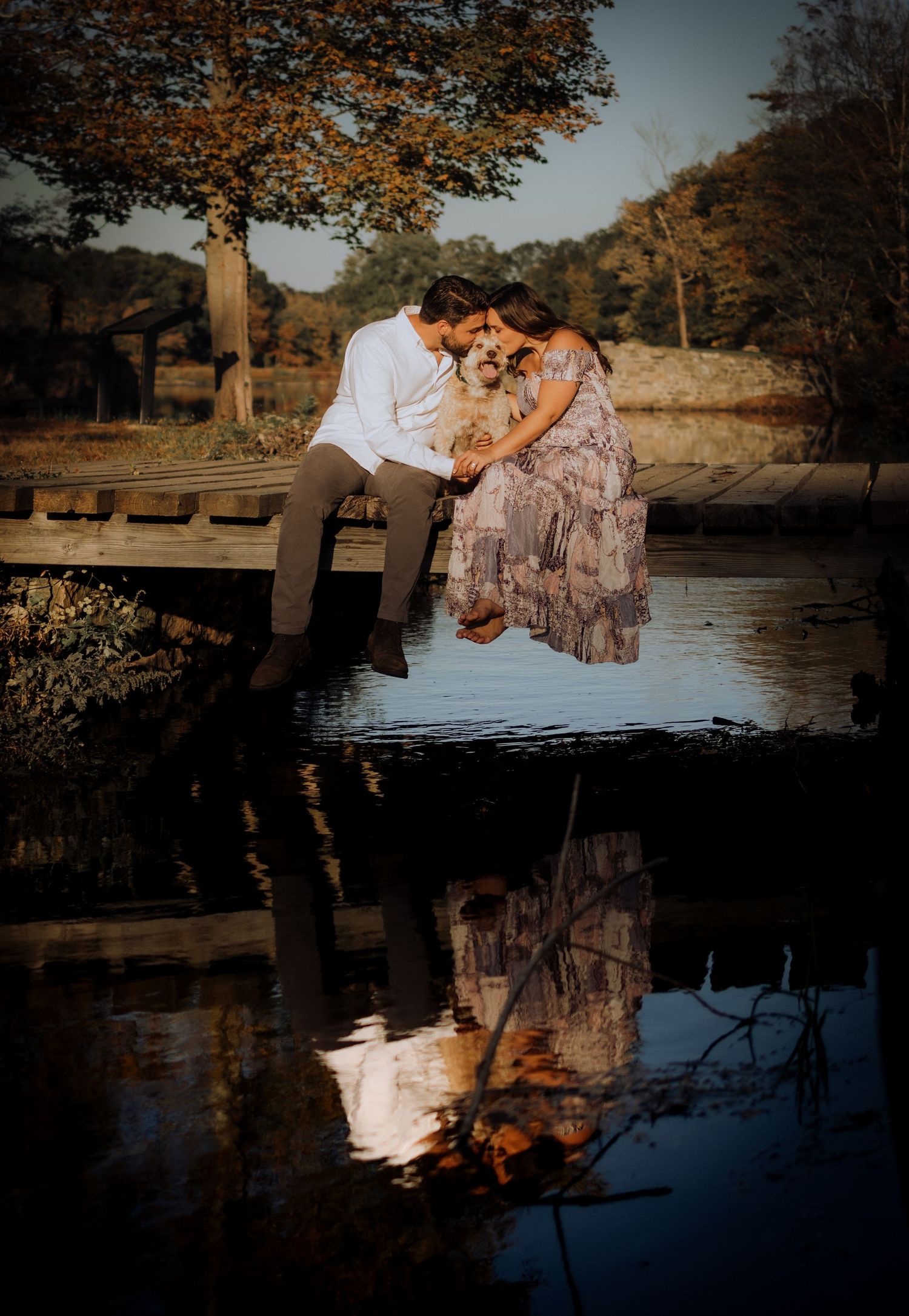 A couple shares a tender moment on a wooden dock overlooking a tranquil lake at sunset.