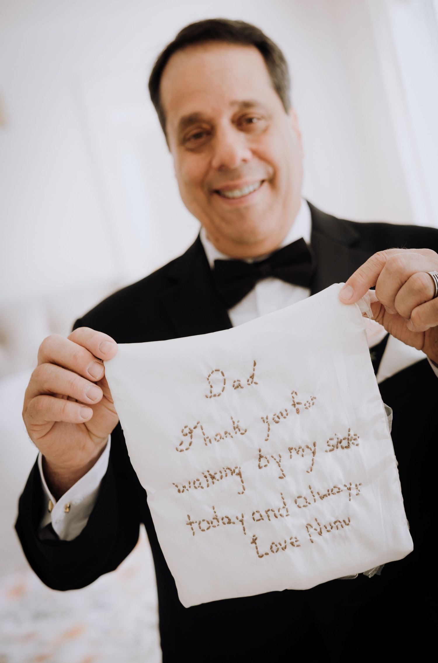 Father of the bride in tuxedo holds embroidered handkerchief reading Dad thank you for walking by my side today and always Love Nora.