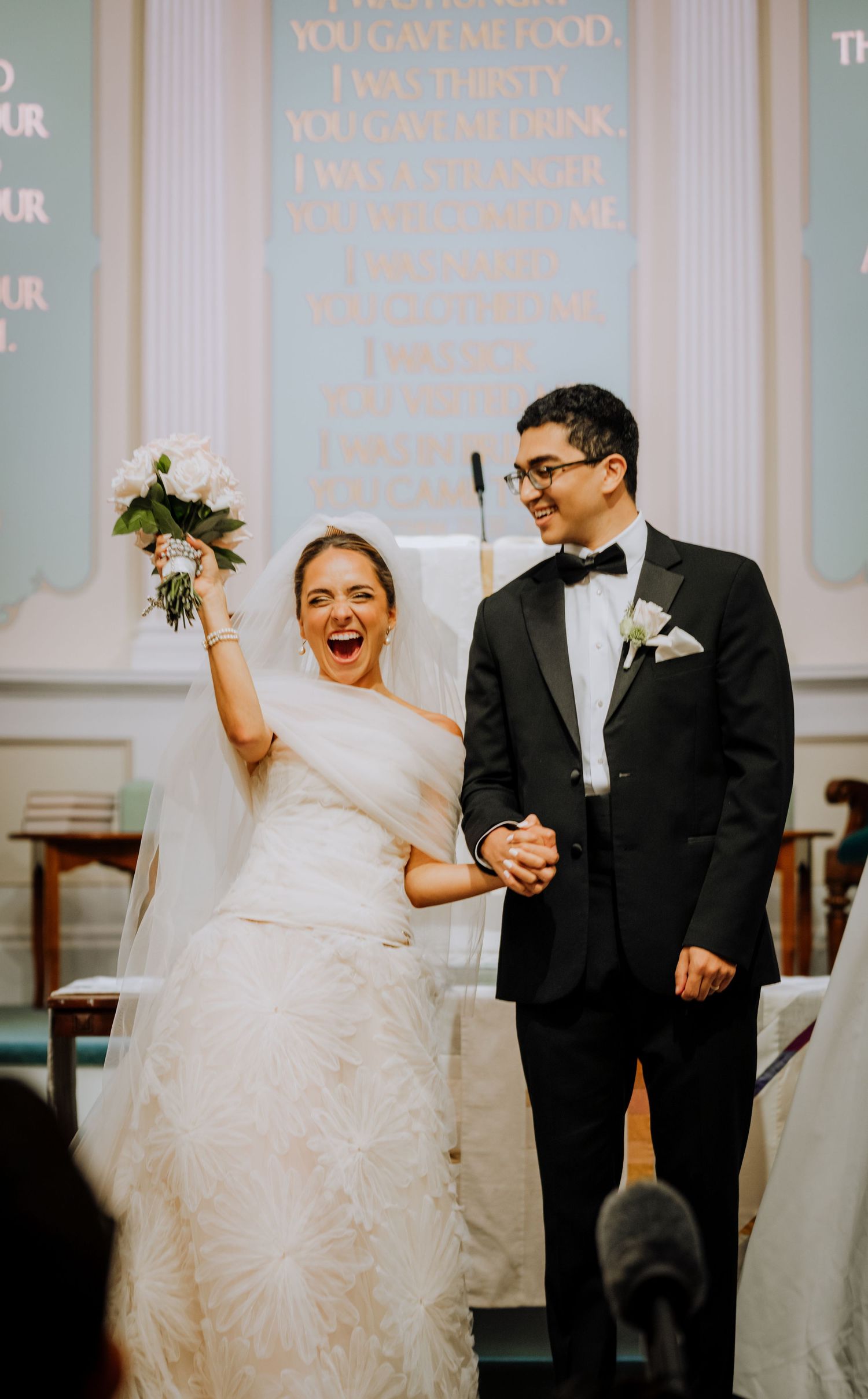 Joyful bride raises bouquet while holding groom's hand at church wedding ceremony altar.