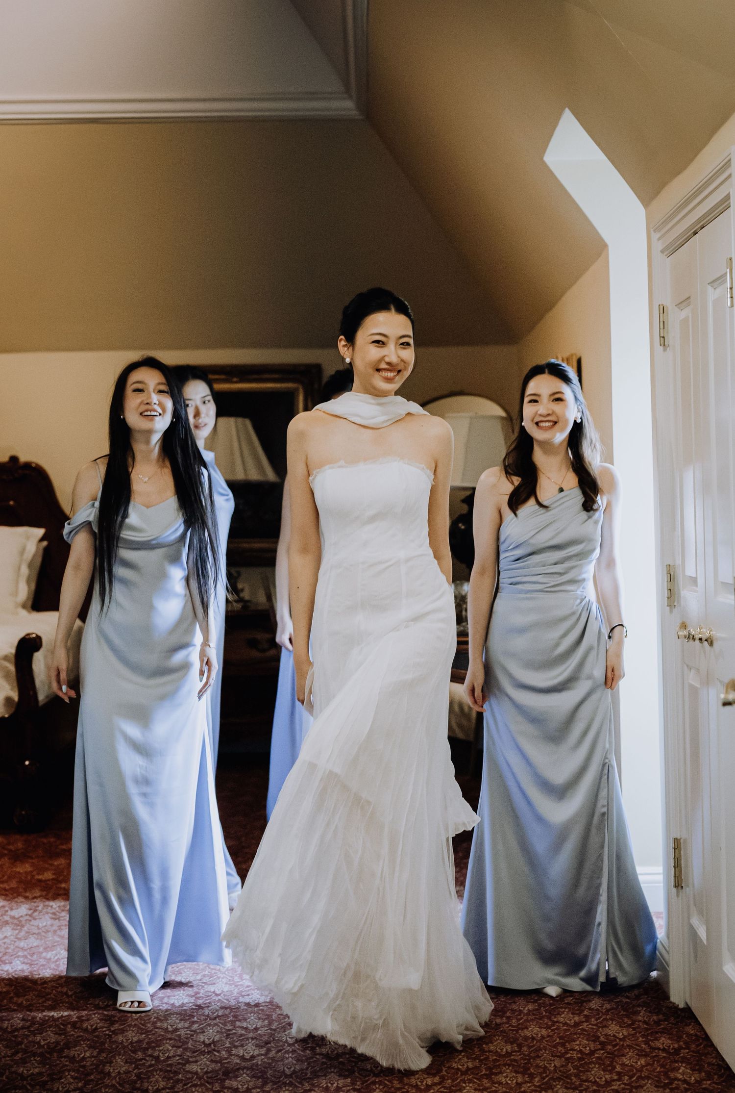 Bride in white mermaid gown smiles with two bridesmaids in light blue satin dresses inside a elegant room.