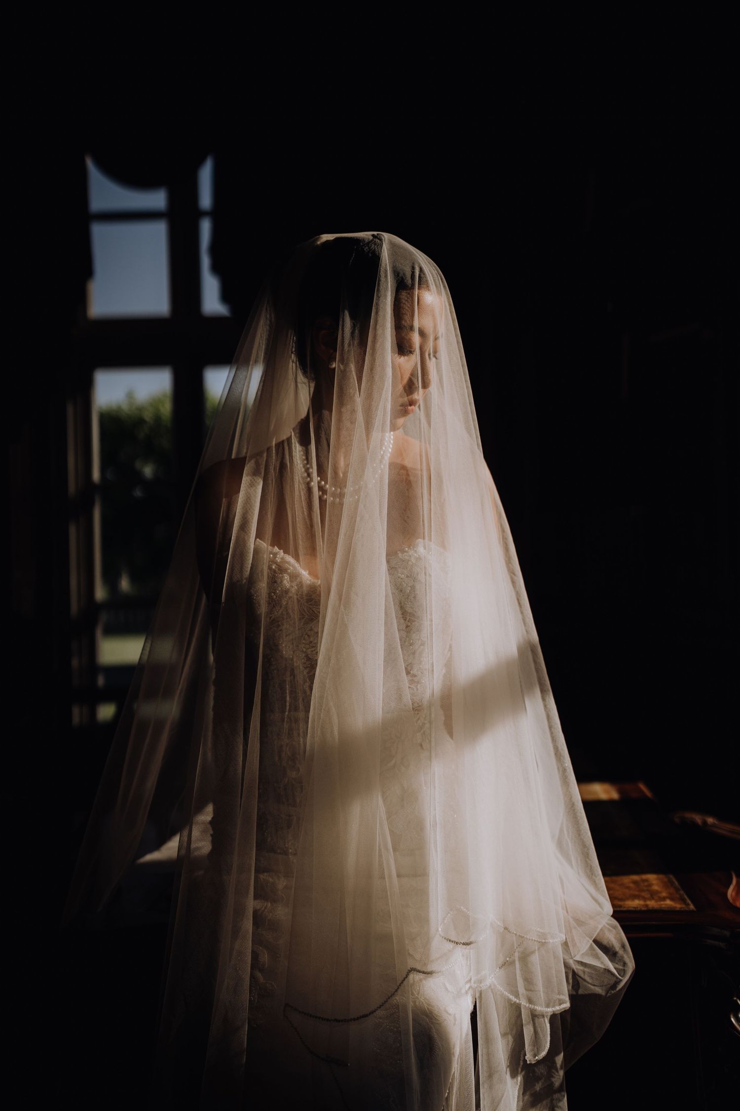 Bride in white wedding dress and veil standing in soft natural light near a dark window.