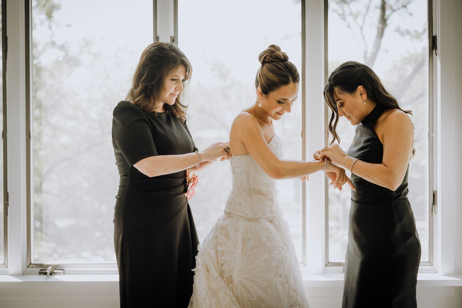 Bride in white lace gown being helped by two bridesmaids in black dresses near bright windows.