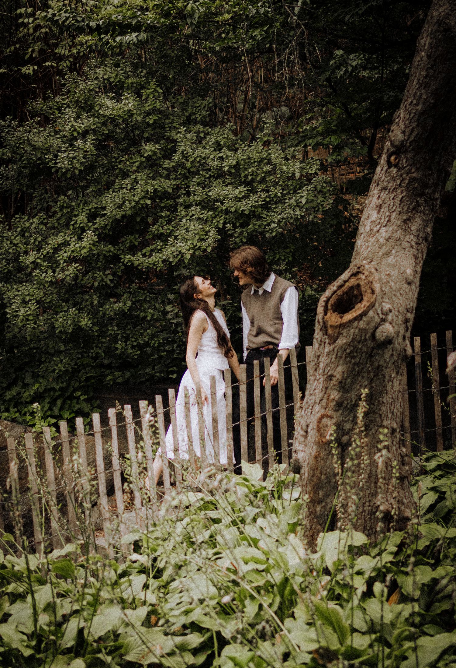 A couple stands together by a rustic wooden fence in a lush forest setting.