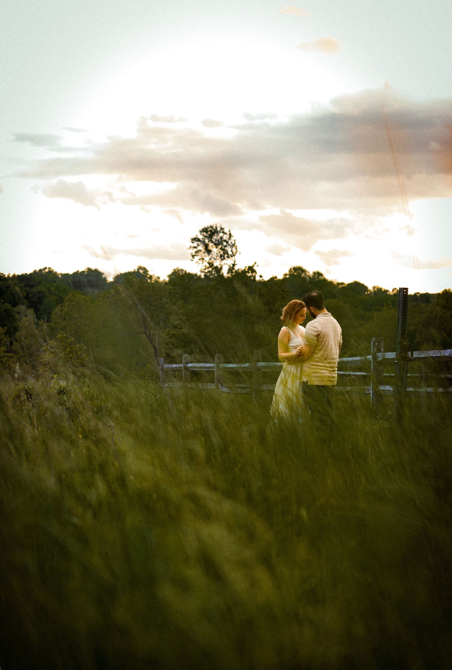 A romantic moment captured in tall grass during golden hour with dramatic sky.