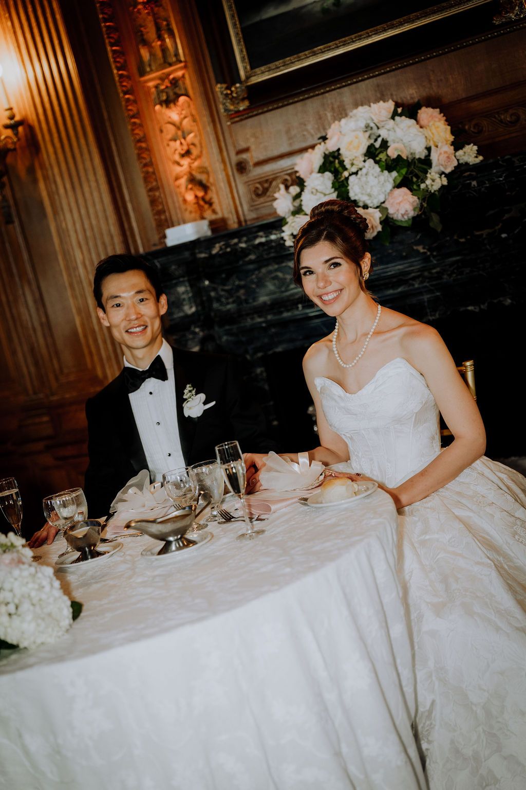Bride and groom smiling at sweetheart table during elegant wedding reception with floral centerpiece.