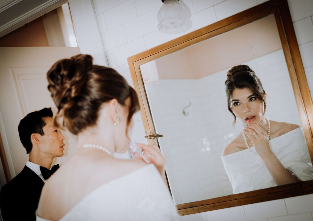 Bride and groom share a tender moment before the mirror, as she adjusts her necklace in a elegant wedding bathroom.