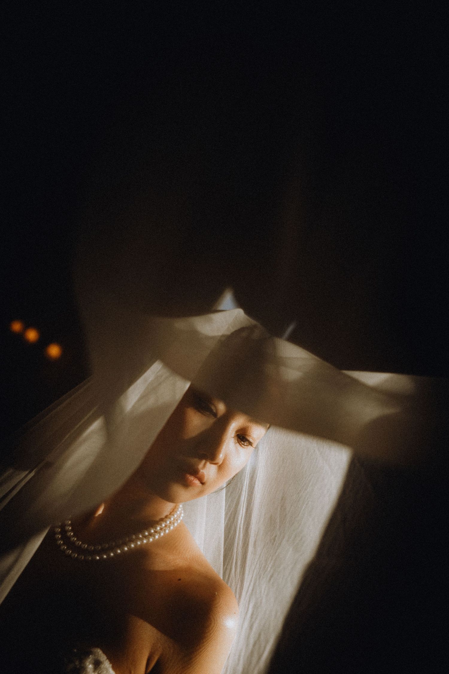 Bride wearing pearl necklace and veil in dramatic moody lighting.