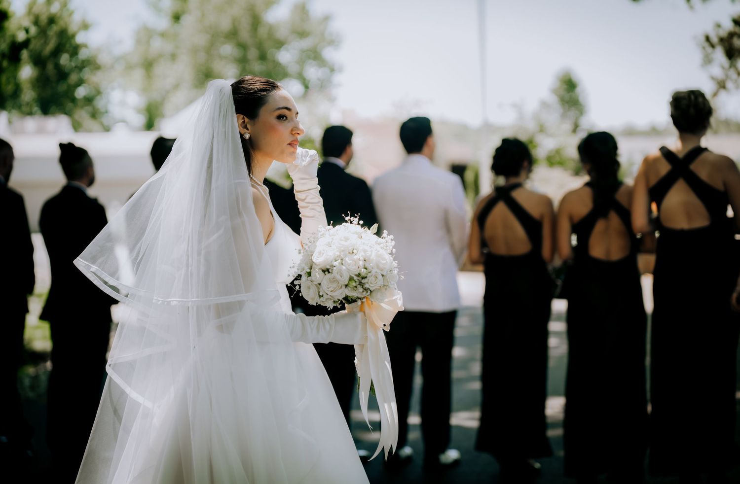 Wedding party lined up outdoors with bridesmaids in black dresses holding white bouquets.