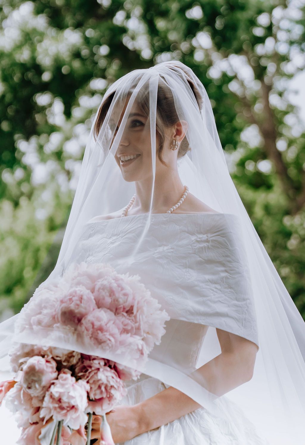 Smiling bride in white gown and veil holding pink peony bouquet outdoors with blooming trees.